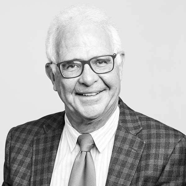 Professional monochrome portrait of a distinguished senior gentleman with white hair, sported with eyeglasses, a plaid-patterned blazer, and tie at our law firm located in Hamilton, Ontario. His countenance exudes warmth and congeniality through his smile.